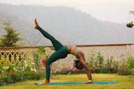 A person is practicing yoga outdoors on a grassy area. They are performing a backbend with one leg raised, on a blue yoga mat. The setting is serene, surrounded by decorative metal railings, flowers, and distant hills in a misty atmosphere.