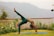 A person is practicing yoga outdoors on a grassy area. They are performing a backbend with one leg raised, on a blue yoga mat. The setting is serene, surrounded by decorative metal railings, flowers, and distant hills in a misty atmosphere.