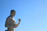A stone statue of a muscular male figure is gazing intently at his clenched fist, set against a clear blue sky.