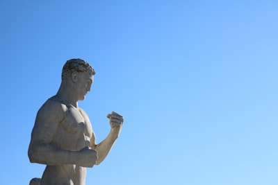 A stone statue of a muscular male figure is gazing intently at his clenched fist, set against a clear blue sky.