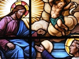 A stained glass artwork depicting religious figures. A bearded man with a halo and robe, symbolizing divinity, is reaching out. A young, winged cherub is positioned on clouds with an arm extended, showing a serene and spiritual moment. Sunrays and luminous backgrounds enhance the divine presence, with vivid colors highlighting the figures.