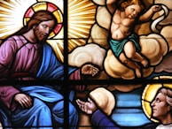 A stained glass artwork depicting religious figures. A bearded man with a halo and robe, symbolizing divinity, is reaching out. A young, winged cherub is positioned on clouds with an arm extended, showing a serene and spiritual moment. Sunrays and luminous backgrounds enhance the divine presence, with vivid colors highlighting the figures.