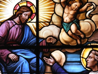 A stained glass artwork depicting religious figures. A bearded man with a halo and robe, symbolizing divinity, is reaching out. A young, winged cherub is positioned on clouds with an arm extended, showing a serene and spiritual moment. Sunrays and luminous backgrounds enhance the divine presence, with vivid colors highlighting the figures.