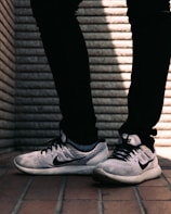 A pair of white and black sneakers with a prominent logo on the side is worn by a person dressed in black pants. The background consists of a textured brick wall with distinct horizontal lines, and the floor is made up of reddish-brown tiles.