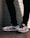 A pair of white and black sneakers with a prominent logo on the side is worn by a person dressed in black pants. The background consists of a textured brick wall with distinct horizontal lines, and the floor is made up of reddish-brown tiles.