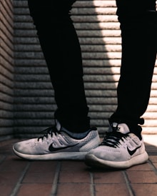 A pair of white and black sneakers with a prominent logo on the side is worn by a person dressed in black pants. The background consists of a textured brick wall with distinct horizontal lines, and the floor is made up of reddish-brown tiles.