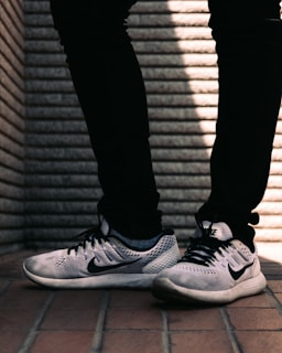 A pair of white and black sneakers with a prominent logo on the side is worn by a person dressed in black pants. The background consists of a textured brick wall with distinct horizontal lines, and the floor is made up of reddish-brown tiles.