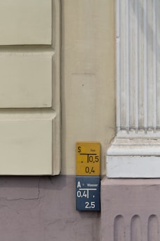 Wall-mounted utility markers with detailed numerical information related to gas and water lines. The yellow sign at the top indicates gas, displaying numbers '10.5' and '0.4'. Below it, a blue sign labeled 'Wasser' provides the figures '0.41' and '2.5'. The markers are affixed to a wall with muted tones of cream and light purple, next to architectural molding.