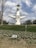 A large, artistic sculpture with a futuristic design featuring a face in its center and extending arms is situated in a grassy area. There are small white flowers in front of the sculpture, with a bare tree on the left side. The sculpture has a circular face at the top with binocular-like eyes. The sky is partly cloudy.