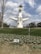 A large, artistic sculpture with a futuristic design featuring a face in its center and extending arms is situated in a grassy area. There are small white flowers in front of the sculpture, with a bare tree on the left side. The sculpture has a circular face at the top with binocular-like eyes. The sky is partly cloudy.