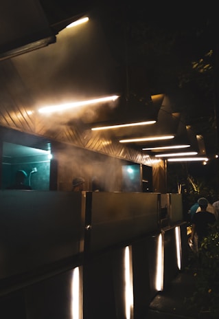 The image depicts a modern, industrial kitchen area at night with bright strip lights on the ceiling. Steam is visible rising, indicating active cooking. Several individuals wearing hats or hair coverings are working in the kitchen. The overall scene has a somewhat futuristic look due to the lighting and metallic surfaces.