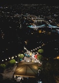 A rooftop terrace at night overlooking a cityscape with lit bridges and buildings reflected in a river. Several people are gathered under string lights, sitting and standing around tables on the terrace, creating a cozy, social atmosphere. Lush greenery surrounds the area, enhancing the intimate setting.