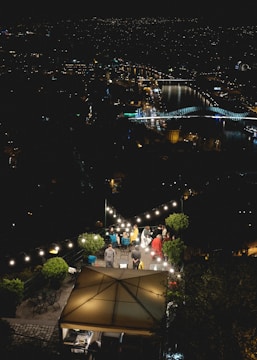 A rooftop terrace at night overlooking a cityscape with lit bridges and buildings reflected in a river. Several people are gathered under string lights, sitting and standing around tables on the terrace, creating a cozy, social atmosphere. Lush greenery surrounds the area, enhancing the intimate setting.