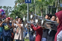 A group of people are playing brass instruments in an outdoor setting. They are lined up side by side, suggesting a band or musical performance. Various individuals are focused on their instruments, surrounded by lush, green palm trees indicating a warm climate. In the background, a colorful balloon stand is partially visible.