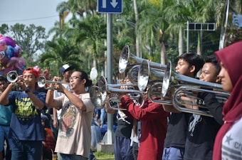 A group of people are playing brass instruments in an outdoor setting. They are lined up side by side, suggesting a band or musical performance. Various individuals are focused on their instruments, surrounded by lush, green palm trees indicating a warm climate. In the background, a colorful balloon stand is partially visible.