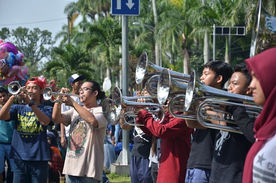 A group of people are playing brass instruments in an outdoor setting. They are lined up side by side, suggesting a band or musical performance. Various individuals are focused on their instruments, surrounded by lush, green palm trees indicating a warm climate. In the background, a colorful balloon stand is partially visible.