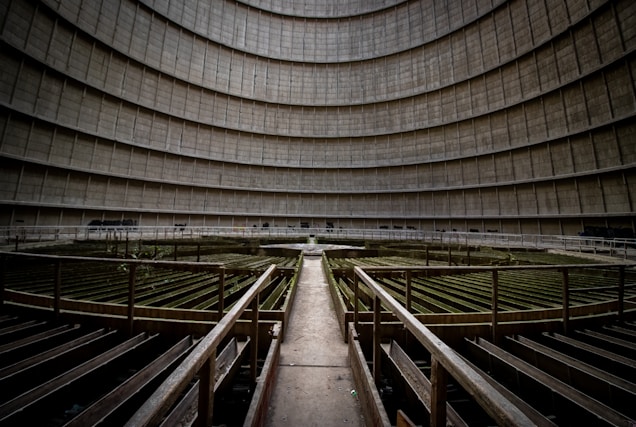 An abandoned industrial site with a large, circular cooling tower interior. The structure's walls curve upwards, creating a sense of emptiness and vastness. Metal walkways and platforms, covered with moss and rust, crisscross the floor, leading towards the center of the space.