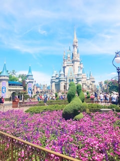 A grand castle stands tall in the background, with intricate spires and detailed architecture. In the foreground, vibrant flower gardens are visible, featuring deep purple blooms bordered by well-maintained hedges and topiary sculptures. A clear blue sky extends overhead, adding a bright and lively atmosphere. Several people are walking around, enjoying the scenic surroundings and lively environment.