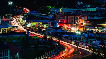 A vibrant cityscape at night with brightly illuminated streets and buildings. Light trails from vehicles create dynamic streaks across the roads. The central area features colorful signage and bustling activity. The scene is accentuated by the vibrant lights and the darkness of the evening sky.