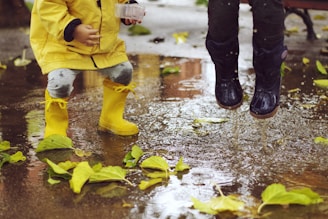 Children wearing rain boots are playing in a puddle. One child is dressed in a yellow raincoat and matching yellow boots, while the other wears dark rain boots. Green leaves are scattered on the wet ground, and water splashes as they jump.