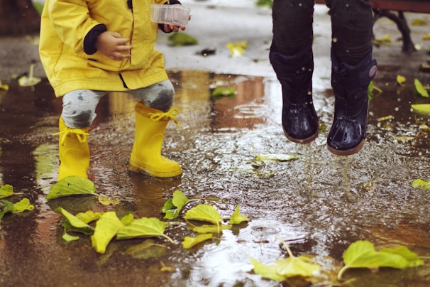 Children wearing rain boots are playing in a puddle. One child is dressed in a yellow raincoat and matching yellow boots, while the other wears dark rain boots. Green leaves are scattered on the wet ground, and water splashes as they jump.