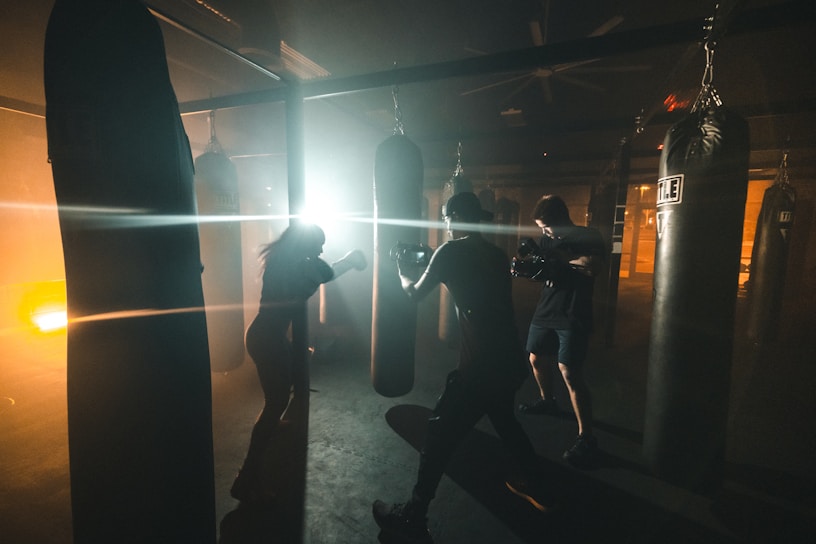 In a dimly lit gym, two people are engaged in boxing practice with punching bags. The area is illuminated by bright artificial lights, creating dramatic contrasts between shadows and light. The atmosphere appears intense and focused, with the individuals fully immersed in their workout.