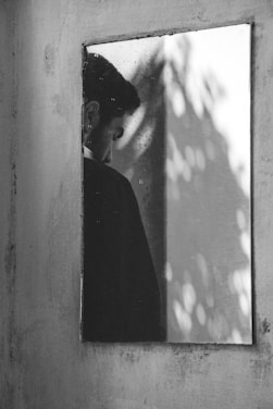 A reflective surface captures the image of a person, positioned near the left side of the frame, looking downward with a solemn expression. The background exhibits a blurred effect with light patches and shadows, creating an abstract play of light and shade.
