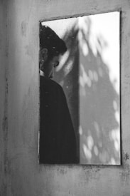 A reflective surface captures the image of a person, positioned near the left side of the frame, looking downward with a solemn expression. The background exhibits a blurred effect with light patches and shadows, creating an abstract play of light and shade.