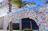 A large cruise ship is docked near a decorated entrance arch, covered with numerous colorful stickers and decals, with palm trees nearby. The arch features playful and vibrant colors, creating a festive and lively atmosphere.