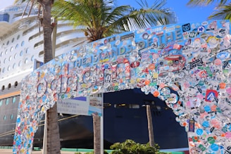 A large cruise ship is docked near a decorated entrance arch, covered with numerous colorful stickers and decals, with palm trees nearby. The arch features playful and vibrant colors, creating a festive and lively atmosphere.