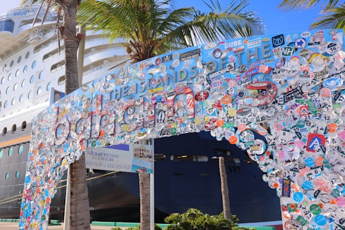 A large cruise ship is docked near a decorated entrance arch, covered with numerous colorful stickers and decals, with palm trees nearby. The arch features playful and vibrant colors, creating a festive and lively atmosphere.