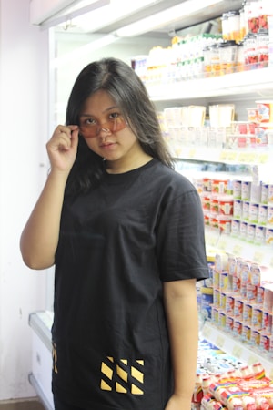 A person with long dark hair wearing a black shirt and holding sunglasses down slightly in a grocery store aisle. A variety of products, including cartons and bottles, are displayed on the shelves behind them, suggesting a chilled drinks or dairy section.