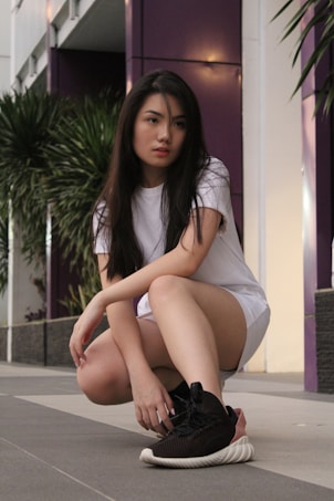A person with long dark hair wearing a white t-shirt and shorts is squatting on a paved pathway. In the background, there are purple pillars and green plants against a white building facade.