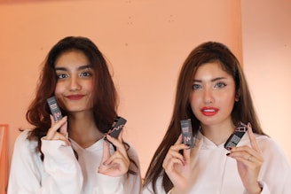 Two individuals are holding and displaying matte lipstick products. They are both wearing white hoodies and have a warm-colored backdrop.