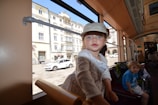 A child wearing a cap leans towards the camera inside a tram. Another child is seated further back, next to a stroller. The tram's interior features wood paneling. Through the window, a city street with historic buildings and a parked white car is visible.