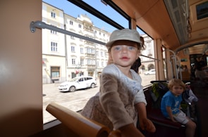 A child wearing a cap leans towards the camera inside a tram. Another child is seated further back, next to a stroller. The tram's interior features wood paneling. Through the window, a city street with historic buildings and a parked white car is visible.