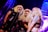 Three individuals with long, blonde wigs and makeup are gathered around a vintage-style microphone, possibly performing on stage. They are dressed in edgy, dark clothing, including leather-like materials and fishnets. The backdrop is a swirl of vibrant blues and purples, adding a theatrical atmosphere.
