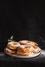 A stack of freshly baked pastries is arranged on a plate, lightly dusted with powdered sugar. The pastries have a golden-brown crispy texture and are highlighted against a dark background, creating a dramatic and appetizing look. A hint of green foliage and a slice of lemon can be seen in the background.