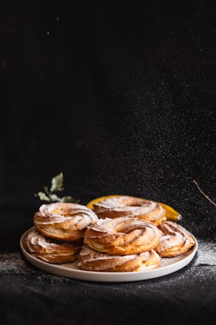 A stack of freshly baked pastries is arranged on a plate, lightly dusted with powdered sugar. The pastries have a golden-brown crispy texture and are highlighted against a dark background, creating a dramatic and appetizing look. A hint of green foliage and a slice of lemon can be seen in the background.
