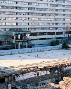 A large, multi-story building with numerous identical windows and air conditioning units. The foreground shows a damaged or partially demolished area with rubble. A person wearing a red helmet is pushing a wheelbarrow across an empty, flat surface.