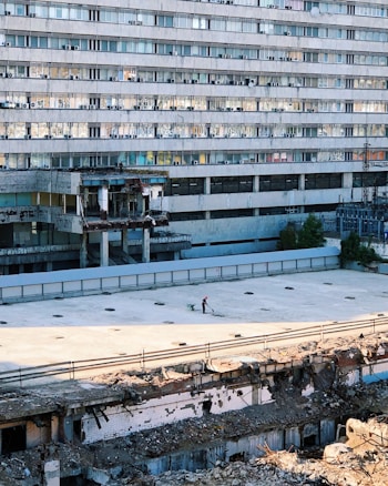 A large, multi-story building with numerous identical windows and air conditioning units. The foreground shows a damaged or partially demolished area with rubble. A person wearing a red helmet is pushing a wheelbarrow across an empty, flat surface.