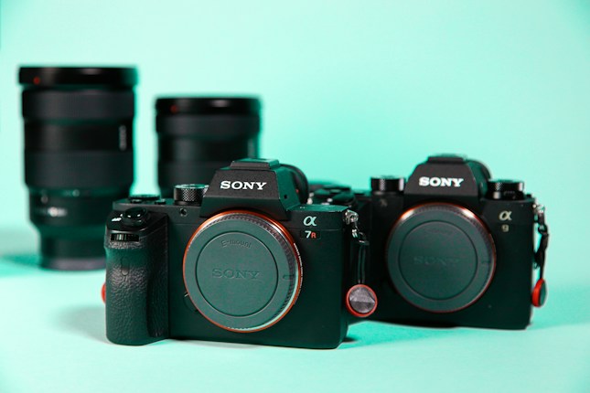 Two Sony Alpha mirrorless cameras are positioned in the foreground, with interchangeable lenses placed in the background. The cameras and lenses are placed on a light turquoise surface, which contrasts with their black bodies.