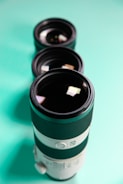 Three camera lenses are arranged in a vertical line on a turquoise surface, with the largest lens in the foreground. The lenses feature black and white exteriors with intricate detailing.