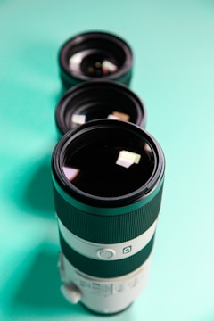Three camera lenses are arranged in a vertical line on a turquoise surface, with the largest lens in the foreground. The lenses feature black and white exteriors with intricate detailing.