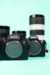 Two Sony cameras are placed side by side with interchangeable lenses in the background against a mint green backdrop. The cameras are prominently displayed in the foreground showcasing their branding and are complemented by a blurred view of two camera lenses.