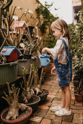 A young child wearing a floral shirt and denim overalls is holding a blue watering can, tending to potted plants arranged on brick paving. Surrounding the child are various plants, some inside pots, along with small garden decorations including miniature houses.