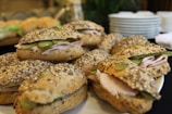A collection of seeded sandwich rolls filled with slices of meat and pickles arranged on a serving platter. In the background, there are stacked plates and a blurred green leafy plant, suggesting a buffet or catering setting.