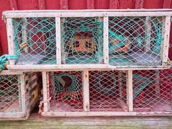 A stack of wooden lobster traps, featuring a grid of turquoise and red netting against a textured red wall. The traps are weathered, indicating they have been used multiple times, and they are interwoven with ropes of various colors.