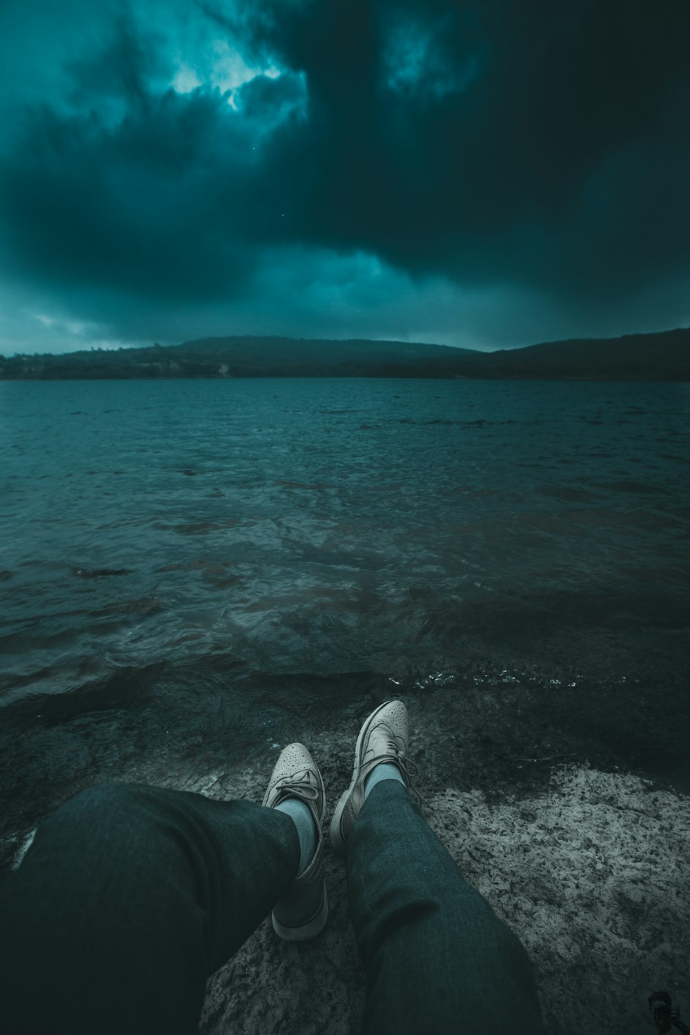 person sitting on rock facing body of water under gray clouds