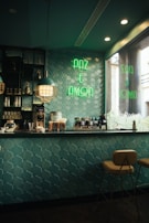 A cozy café interior with teal geometric wall tiles and a countertop equipped with coffee machines and various supplies. The space is lit with stylish pendant lights and has a neon sign in green reading 'PAZ E AMOR' on the wall. Two warm-colored stools are positioned at the bar, creating a welcoming atmosphere.